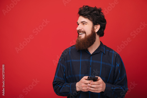 Portrait of bearded guy holding a mobile and looking away, standing over red wall © Vulp