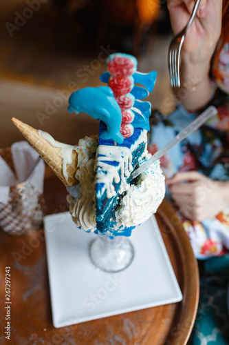 Close-up of crazy giant ice cream in large glass with waffle cone, piece of cake, marmalade, cream, and milk cocktail inside. Insanely delicious and high-calorie sweet ice cream milkshake in cozy cafe