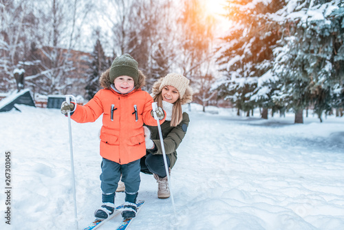 Young mother woman happy smiling, son boy 3 years old laughs. In winter, outside park, background is snow drifts Christmas tree. Free space. Children's skis, skiing lessons, first steps, child care. photo