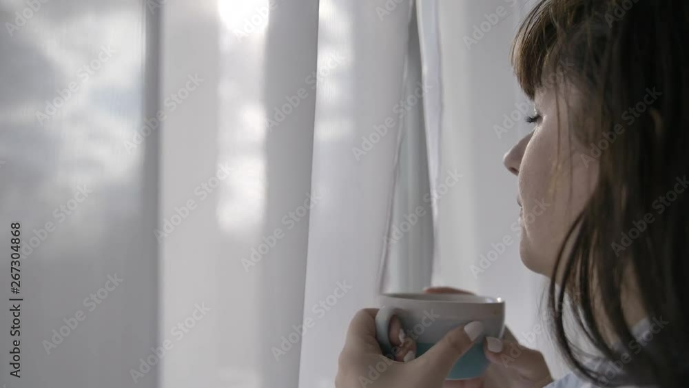 happy woman takes a sip of coffee and enjoys the taste, standing near the window happy woman takes a sip of coffee and enjoys the taste, standing near the window
