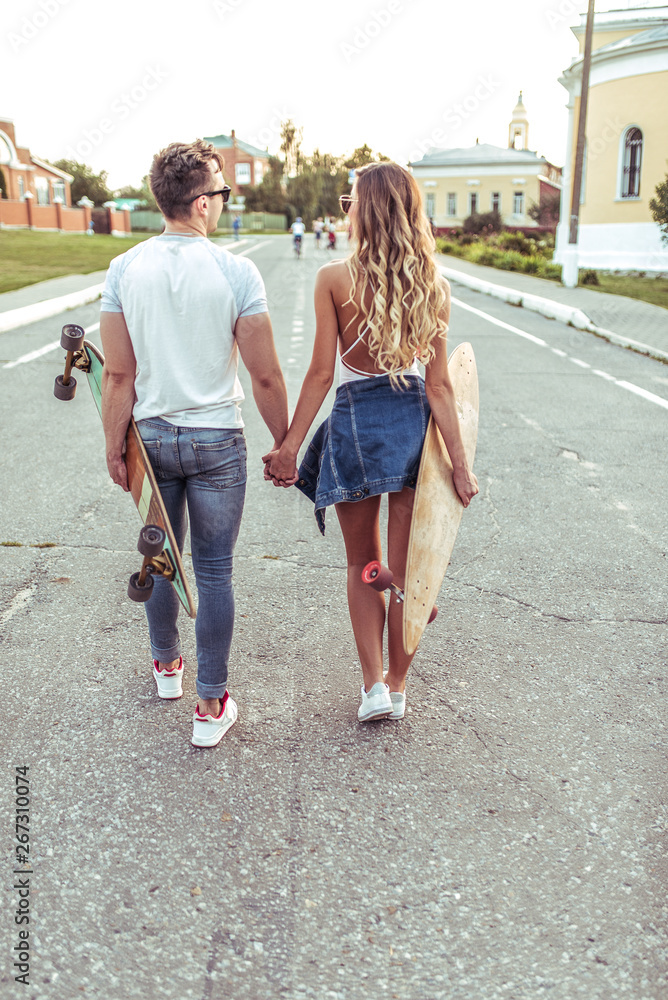 Young couple man woman walk summer city, background road view from rear. In hands skateboard, longboard. Holding hands, concept love, caring, understanding, relationship. Empathy care inspiration.