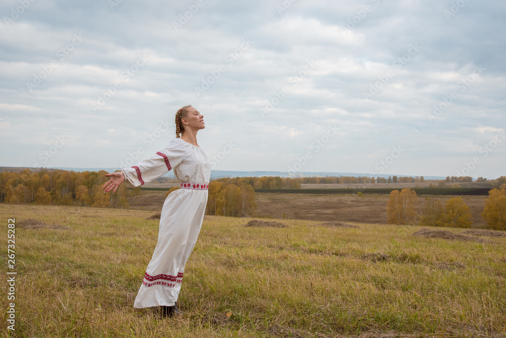 A girl in a Slavic dress with a red ribbon in her hair is standing in a field with her arms open to the sky A girl in a Slavic dress with a red ribbon in her hair is standing in a field with her arms open to the sky