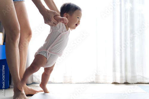 Non focus on moving Asian baby taking first steps walk forward on the soft mat with smile. Happy little baby learning to walk with mother help at home. Baby growth and development concept.
