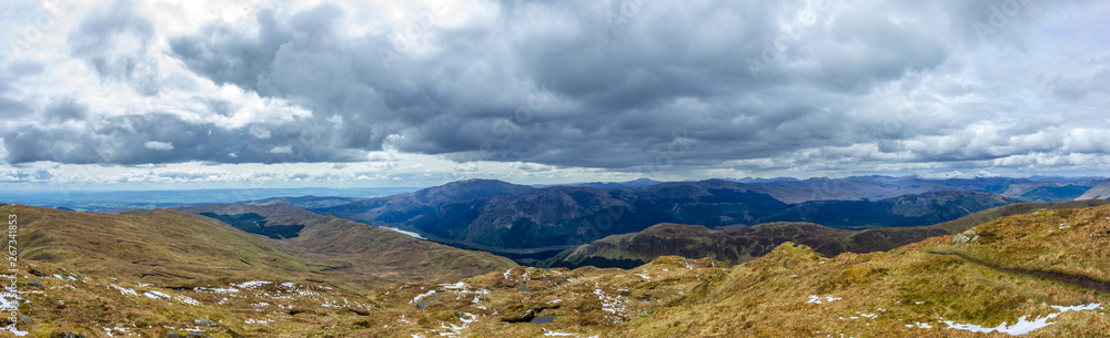 A panorama view of a Scottish mountain range with lakes, pines forest under a majestic cloudy white sky