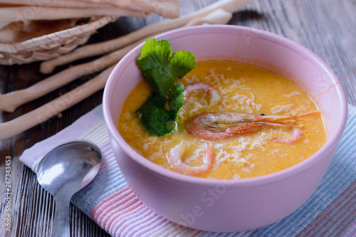 Colorful and tasty cream soup with cheese, shrimp and cilantro leaves on a wooden table next to bread sticks, close-up
