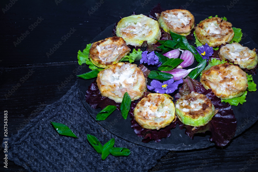 Plakat On a black tray are lettuce leaves, and in them are mugs of roasted zucchini. They are watered with sauce. In the middle are the flowers of violets and garlic cloves.