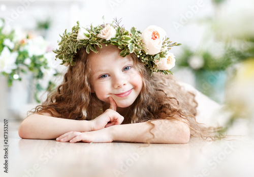 Kid - girl with blond hair in a flower crown. Beautiful little girl in a bright studio lying on the floor and looks into the camera
