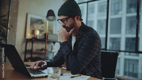 Bearded hipster man wearing eye glasses and using devices at office.Confident busy male freelancer in trendy eyewear using laptop while surfing net searching information for presentation