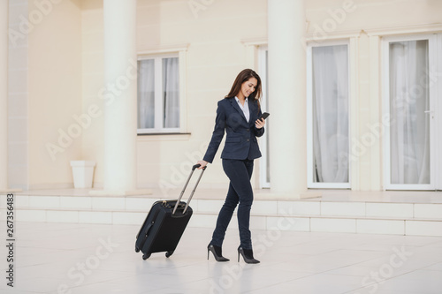 Young smiling Caucasian businesswoman dressed smart casual pulling luggage and using smart phone while walking outdoors. Business trip concept.