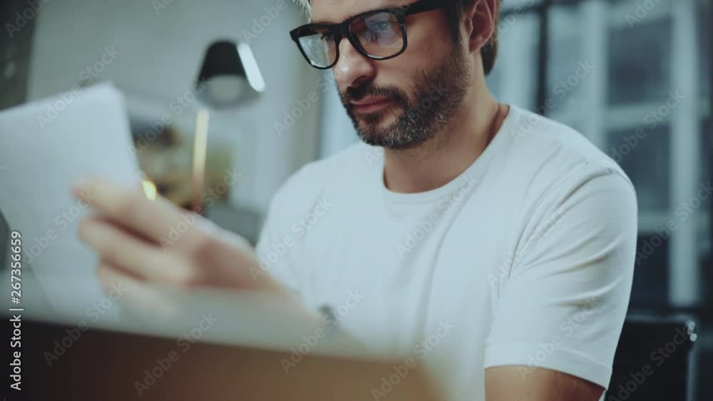Young bearded professional coworker wearing eye glasses working at modern loft studio-office with laptop computer.Big panoranic windows on blurred background Young bearded professional coworker wearing eye glasses working at modern loft studio-office with laptop computer.Big panoranic windows on blurred background