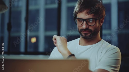 Young bearded professional coworker wearing eye glasses working at modern loft studio-office with laptop computer.Big panoranic windows on blurred background