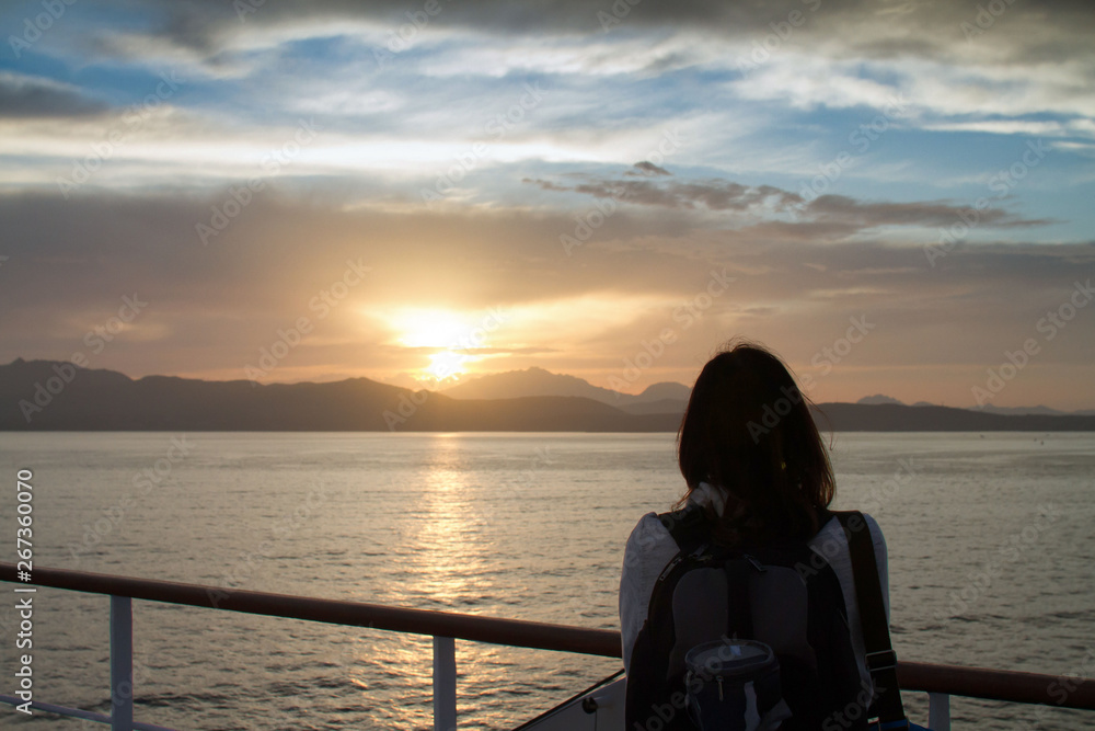 Young woman girl with backpack on shoulder shot from behind admires the sunrise on the Sardinian sea coast with intense orange coloring seen from the sea on the ferry that is about to dock