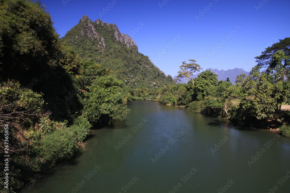 Panoramic scenic view of Nam Song (Xong) river amidst trees and rural karst hills landscape against blue clear sky , near Vang Vieng, Laos Panoramic scenic view of Nam Song (Xong) river amidst trees and rural karst hills landscape against blue clear sky , near Vang Vieng, Laos