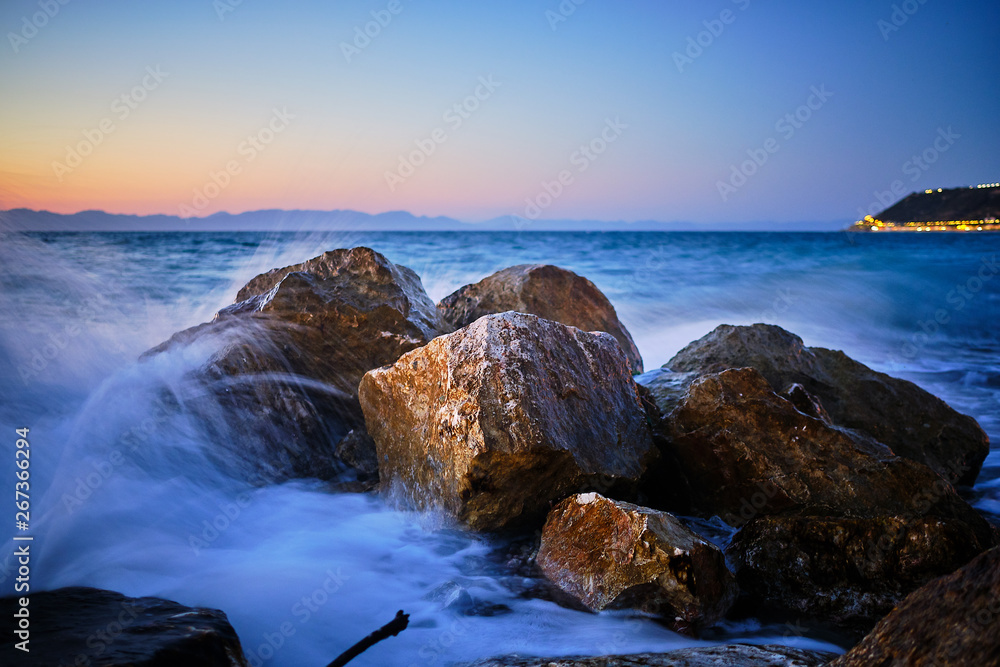 a group of rocks in the foreground with waves foaming below. Street lights in the background