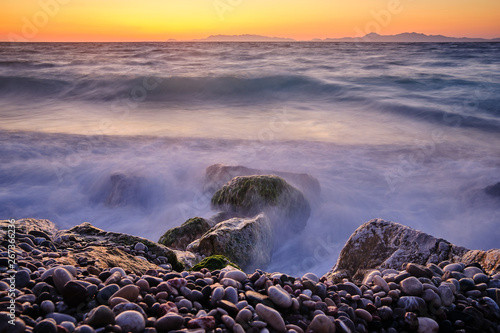 Stone beach in the foreground and large stones with moss in the sea with mountains and sunset in the background