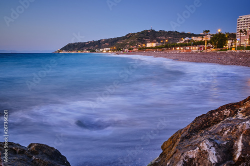 waves sweep in on a beach a summer evening. Big stones in the foreground and hotel in the background photo