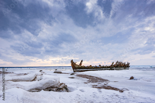 Old broken boat wreck and rocky beach in wintertime. Frozen sea, evening light and icy weather on shore like fairy tale country. Winter on coast. Blue sky, white snow, ice covers the land. photo