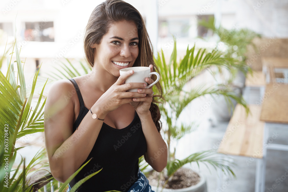 But first coffee. Charming tanned woman enjoy tasty tea hold cup smiling joyfully, start morning contemplating beautiful scenery standing balcony veranda near plants, laughing happily But first coffee. Charming tanned woman enjoy tasty tea hold cup smiling joyfully, start morning contemplating beautiful scenery standing balcony veranda near plants, laughing happily