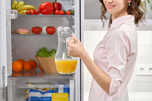 Girl standing refrigerator and holding pitcher with juice.