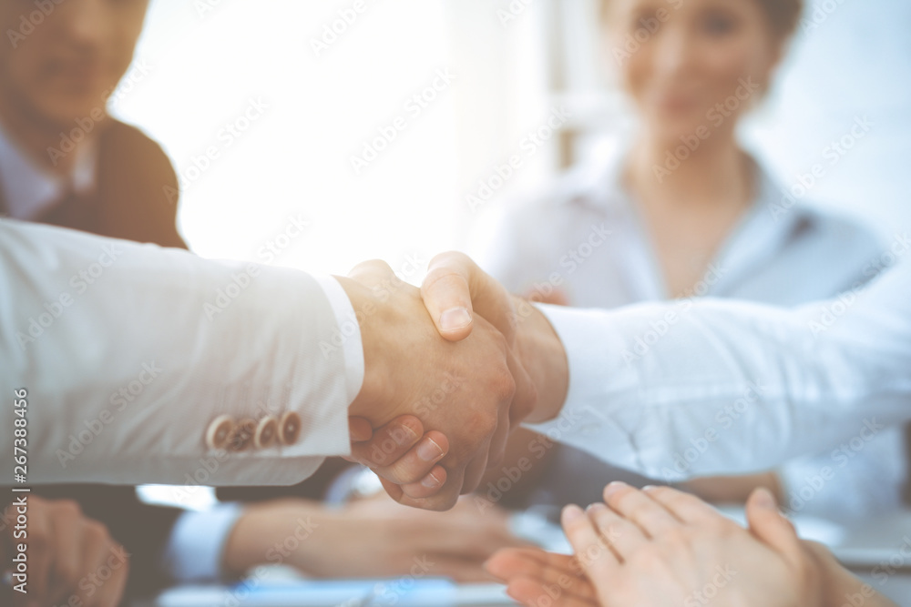 Business people shaking hands at meeting while theirs colleagues clapping and applauding. Group of unknown businessmen and women in modern white office. Success teamwork, partnership and handshake