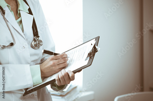 Doctor woman working with medication history records form while standing near window in clinic. Medicine and health care concept. Green is main color