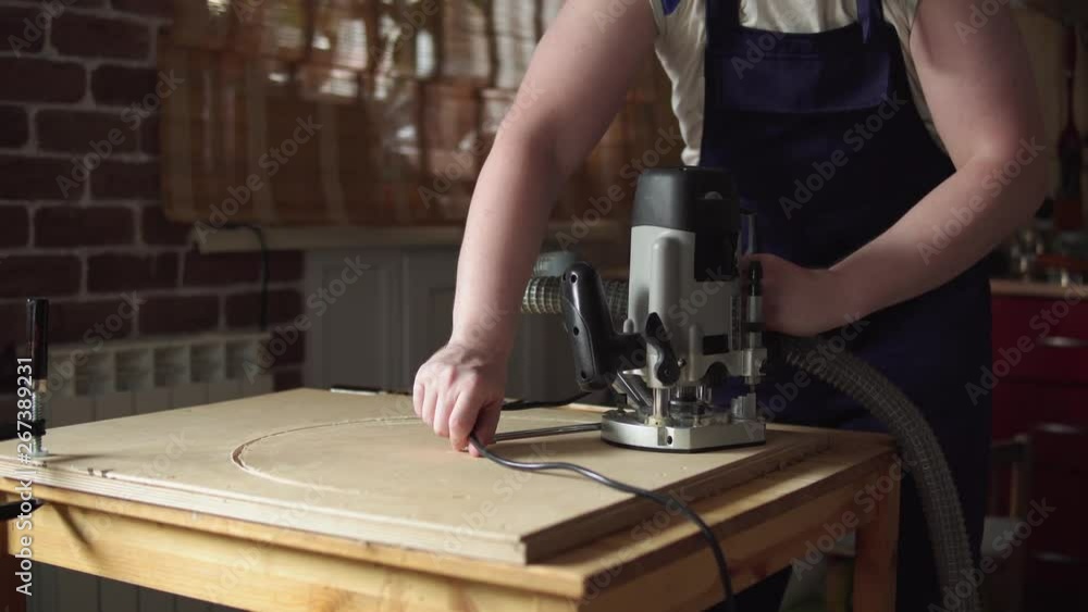 Blond carpenter works with modern hand-held plunge router with dust extraction tube in workshop. Joiner cuts round detail of furniture out of plywood. Blond carpenter works with modern hand-held plunge router with dust extraction tube in workshop. Joiner cuts round detail of furniture out of plywood.