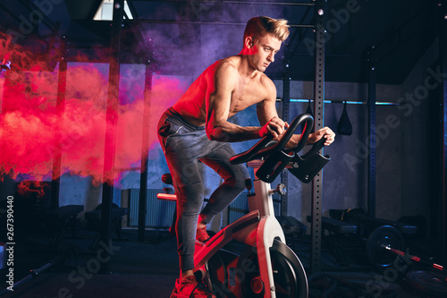 Cardio Workout. Shirtless athletic man training on bicycle in gym. Low key photo of sports guy is exercising on a stationary bike over blue and red smoky background