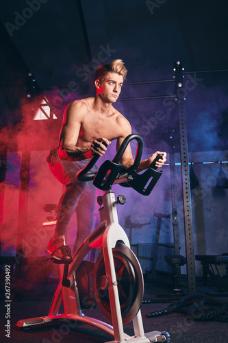 Cardio Workout. Shirtless athletic man training on bicycle in gym. Low key photo of sports guy is exercising on a stationary bike over blue and red smoky background