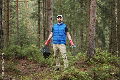Adult man volunteer in red gloves removes plastic trash in forest, caring for environment.