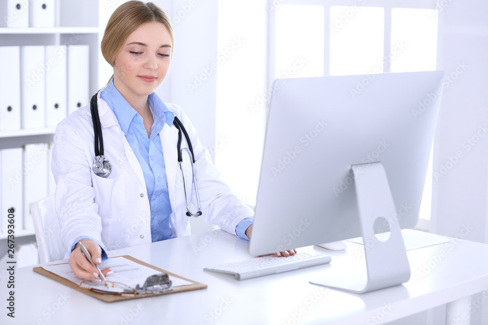 Young woman doctor at work in hospital looking at desktop pc monitor. Physician controls medication history records and exam results. Medicine and healthcare concept Young woman doctor at work in hospital looking at desktop pc monitor. Physician controls medication history records and exam results. Medicine and healthcare concept