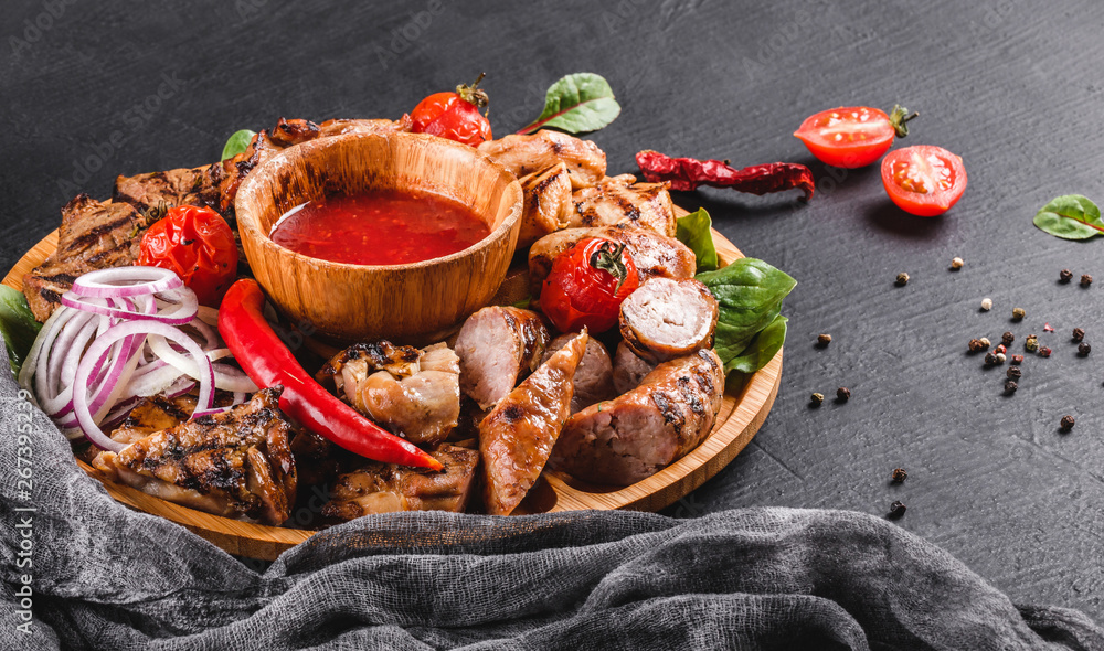 Assorted delicious grilled meat and sausages with tomatoes and bbq sauce on cutting board over black stone background. Hot Meat Dishes. Top view, flat lay