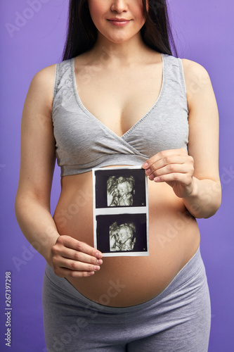 Unrecognizable pregnant white skinned woman in grey home suit with top holding ultrasonic scan in front of her round bare belly, isolated on purple background