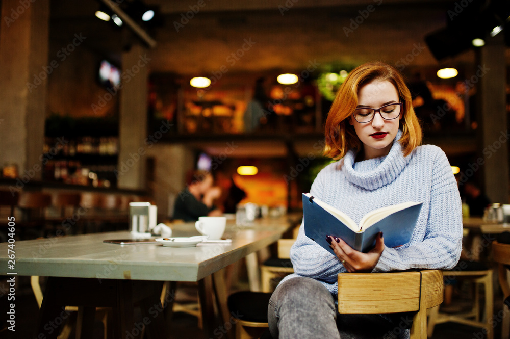 Cheerful young beautiful redhaired woman in glasses sitting at her working place on cafe reading something at her notebook.