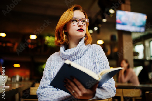 Cheerful young beautiful redhaired woman in glasses sitting at her working place on cafe reading something at her notebook.