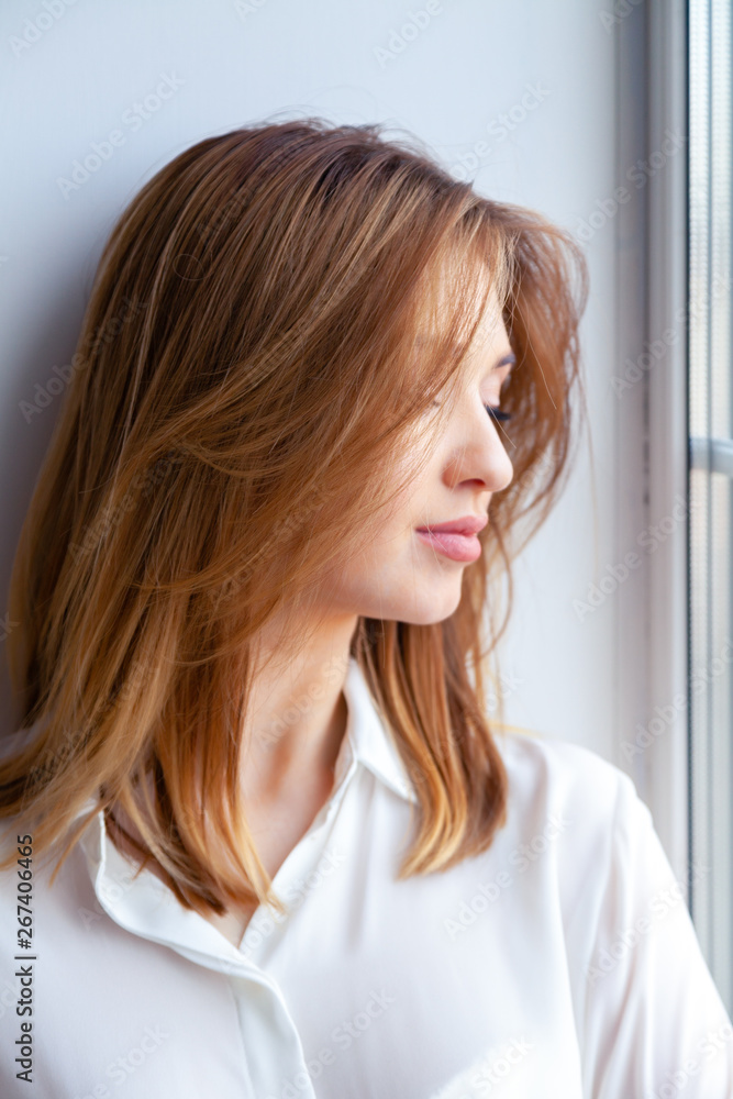 Young beautiful caucasian girl red hair girl in white shirt, blue jeans sitting on window, looking side. Side view, female beauty portrait famous blogger, artist, advert for glance magazine Young beautiful caucasian girl red hair girl in white shirt, blue jeans sitting on window, looking side. Side view, female beauty portrait famous blogger, artist, advert for glance magazine