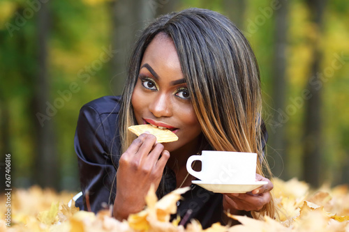 Beautiful young African woman drinking coffee from a white Cup on the background of autumn leaves in the Park
