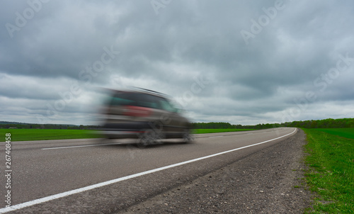 The road through the fields to the horizon and the car in motion at overcast weather