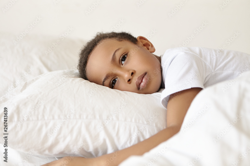 Sadness and loneliness concept. Portrait of sad or thoughtful pensive Afro American male child in white t-shirt lying awake on bed, feeling upset because of nightmare, staring in front of him Sadness and loneliness concept. Portrait of sad or thoughtful pensive Afro American male child in white t-shirt lying awake on bed, feeling upset because of nightmare, staring in front of him