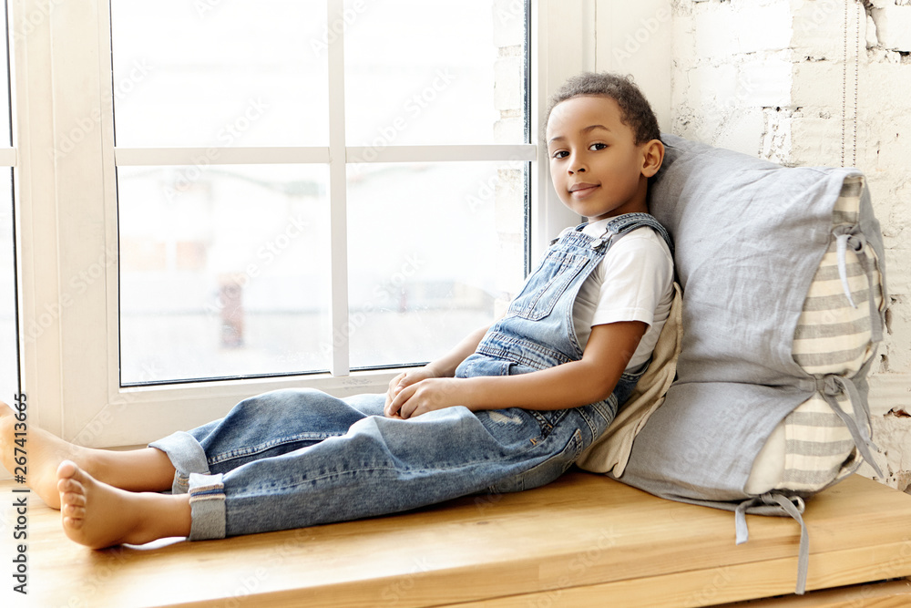 Rest, comfort and relaxation concept. Adorable Afro American black male child in denim jumpsuit sitting barefooted on wooden windowsill, looking outside, having curious interested facial expression