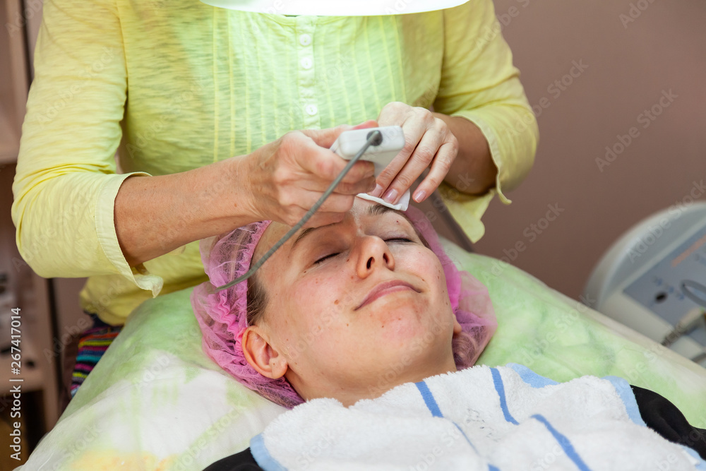 A young girl is lying on couch with a pink hat on her hair while performing an ultrasonic face cleaning with a special device for removing acne and fat deposits under the skin on forehead. Cosmetology