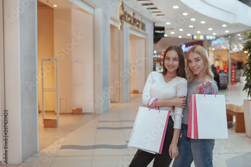Gorgeous young woman smiling to the camera with ehr blond haired female friend, holding shopping bags. Beautiful happy women enjoying shopping together, copy space. Retail, purchasing concept