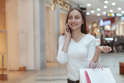 Stunning cheerful female customer walking at the mall, talking on her phone, carrying shopping bags, copy space. Lovely woman calling someone while shopping. News, conenctions concept