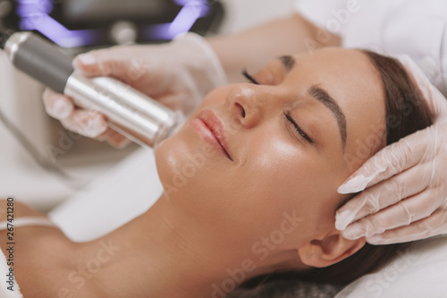 Gorgeous happy young woman with perfect skin smiling with her eyes closed, getting facial treatment at beautician office. Professional cosmetologist performing microcurrent face skin procedure