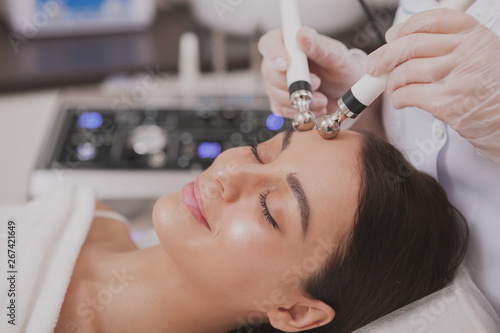 Close up of a lovely relaxed young woman enjoying facial microcurrent therapy by professional cosmetologist. Beautiful woman with perfect skin getting face care procedures by beautician