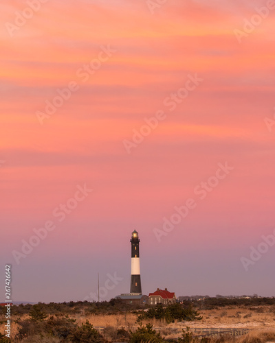 A lighthouse beacon on the horizon under a vibrant pink and orange sunset sky. Fire Island New York. 