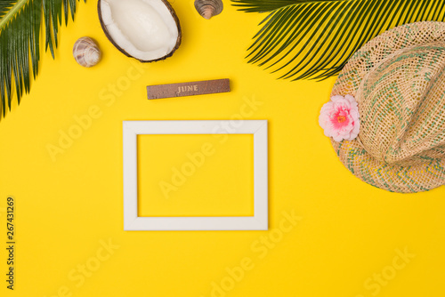 Stylish summer composition with photo frame, green leaves, hat and coconut on a yellow background. Artwork mockup with copy space
