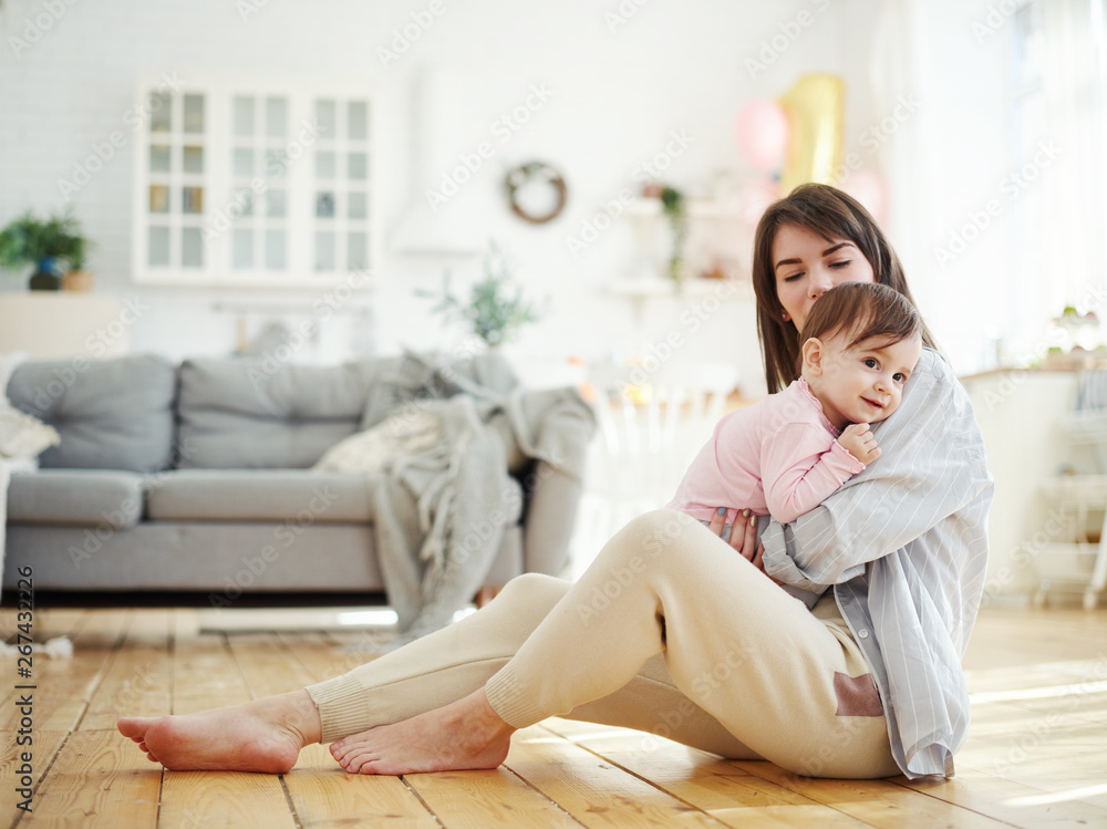 Beautiful young mother sitting on floor in domestic room and hugging her cute smiling baby daughter