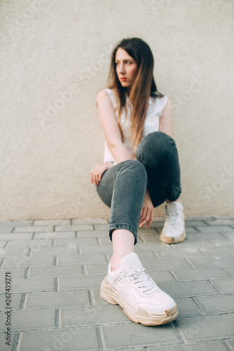 Modern stylish woman in white big sneakers and gray jeans sits resting by the wall. Cool girl resting.
