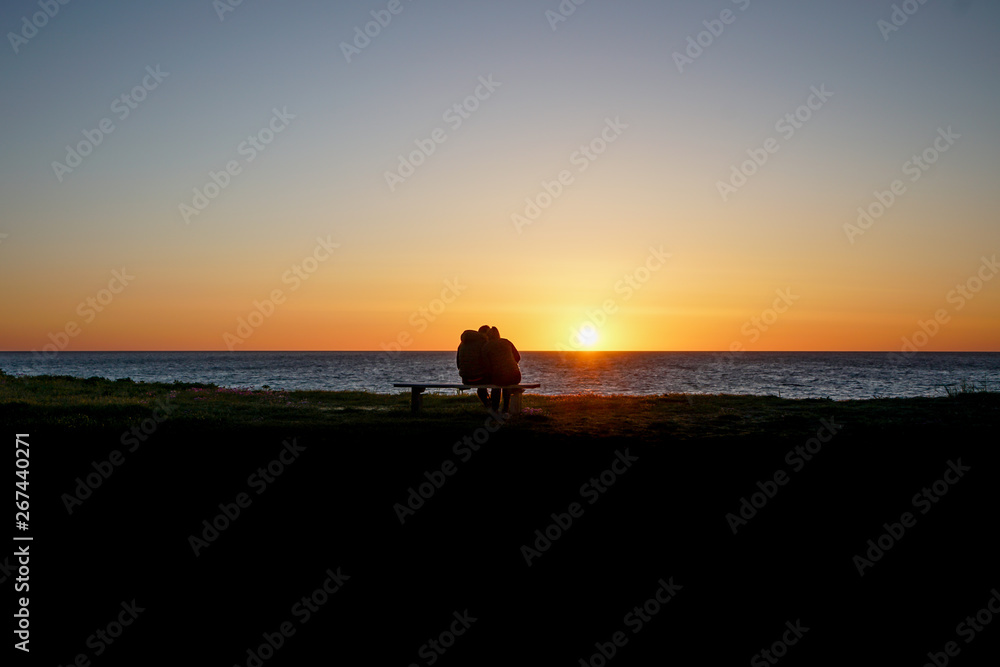 Profile of a young couple sitting on a bench while the sun sets over the ocean.