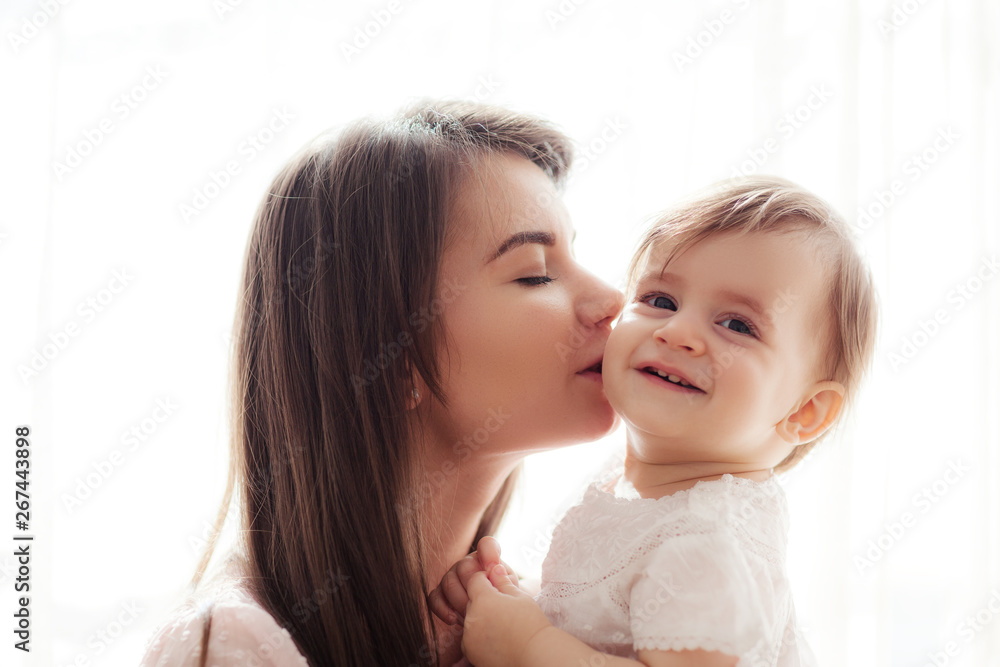 Beautiful portrait of loving mother with closed eyes kissing her happy smiling baby daughter on cheek Beautiful portrait of loving mother with closed eyes kissing her happy smiling baby daughter on cheek